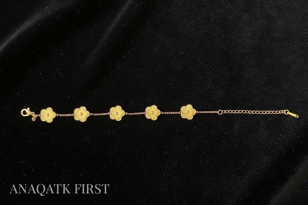 Gold bracelet with floral designs on a black fabric background, featuring the brand 'ANAQATK FIRST'.