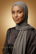 Woman wearing a gray hijab and black top against a dark background with 'ANAQATK FIRST' branding.