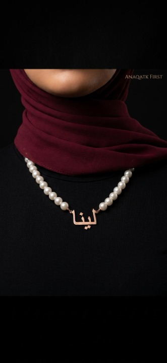 Person wearing a red hijab and pearl necklace with a gold pendant on a black background